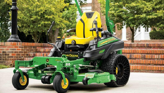 Green and yellow zero-turn lawn mower with black and yellow seat parked on a concrete driveway near brick steps and green trees.