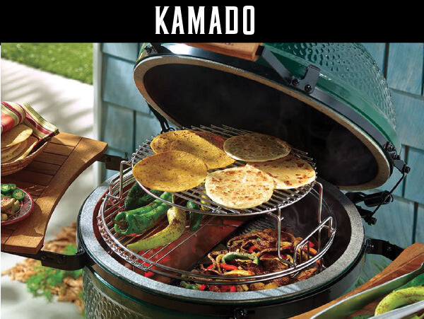 Charcoal kamado grill with its lid open, showing grilled vegetables and flatbreads cooking on the upper and lower racks in an outdoor setting.
