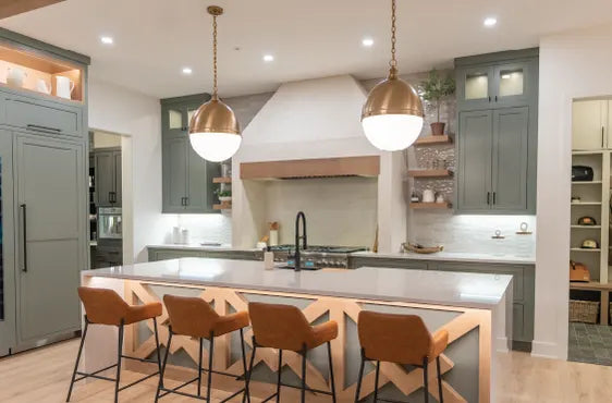 Bright kitchen with a large island featuring a white countertop, four tan leather bar stools, and brass pendant lights above, surrounded by gray cabinetry and open shelves.