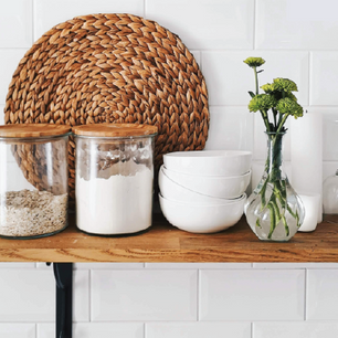 Wooden kitchen shelf with glass jars of dry ingredients, stacked white bowls, a clear vase holding green flowers, and a woven round placemat against white tiled wall.