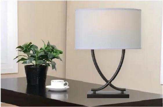 Table lamp with a curved dark metal base and a large white drum shade placed on a polished dark wood surface next to a potted green plant and a white teacup.