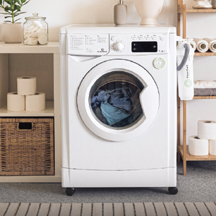 Front-loading white washing machine with clothes inside, positioned between a shelving unit with storage baskets and toilet paper rolls, and a wooden rack holding towels and a handheld vacuum.