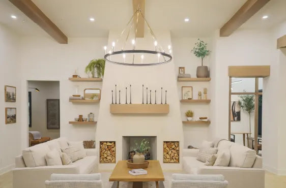 Bright living room interior featuring neutral-toned sofas, wood accents, floating shelves with decor, a modern chandelier, and a central fireplace with stacked firewood.