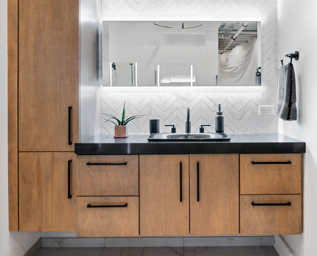 Modern bathroom vanity featuring wood cabinets with black hardware, a black countertop with a sink and faucet, and a large backlit mirror above.