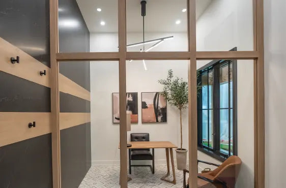 Modern office space with wood-framed glass partitions, a simple wooden desk, black chair, potted plant, and abstract wall art under a contemporary ceiling light.