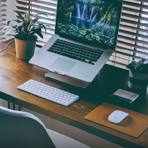 Minimalist wooden desk setup featuring a laptop on a raised stand displaying a waterfall screensaver, wireless keyboard and mouse, two potted plants, and a closed notebook.