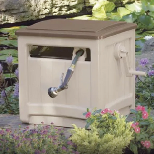 Outdoor beige garden hose storage box with a hand crank on the side, placed among green plants and pink flowers.