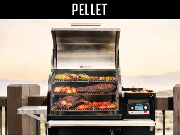 Outdoor pellet grill with its lid open, cooking a variety of meats and vegetables on multiple racks against a scenic background.