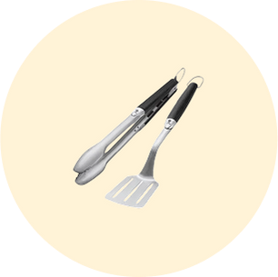 Pair of stainless steel grilling tools with black handles, including tongs and a slotted spatula, on a light beige circular background