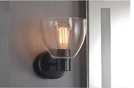 Wall-mounted lighting fixture with a clear glass shade revealing an exposed filament bulb, mounted on a dark metal base against a textured gray wall.