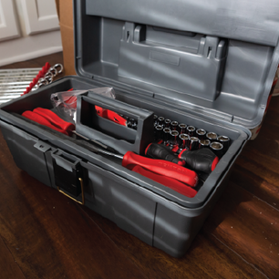 Gray plastic toolbox open on a wooden floor, containing red-handled tools, a set of socket wrenches, and a plastic bag with safety glasses.