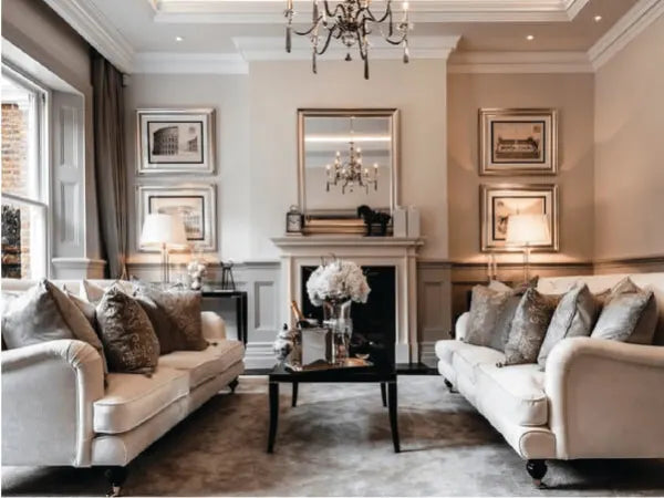 Elegant living room with matching cream sofas adorned with decorative pillows, a black coffee table, ornate chandelier, and classic framed artwork.