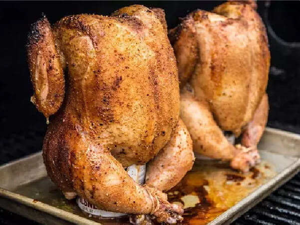 Two whole chickens seasoned and roasted upright on a metal tray with juices collecting beneath them.