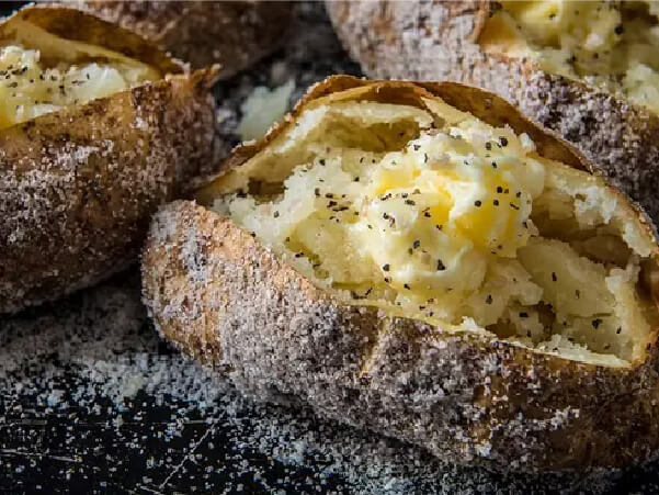Close-up view of baked potatoes with crispy skin, topped with melted butter and freshly cracked black pepper on a dark surface.