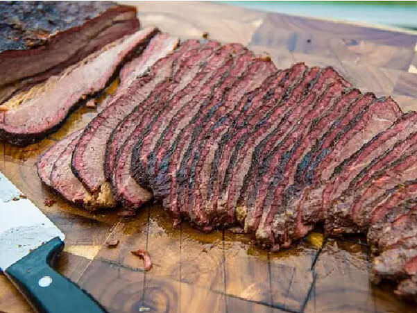 Thin slices of smoked brisket arranged on a wooden cutting board next to a chef’s knife with visible seasoning on the meat’s crust.