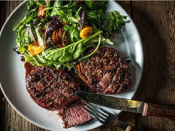 Two grilled filet mignon steaks on a white plate served with mixed greens and cherry tomatoes beside a steak knife and fork.