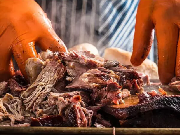 Hands wearing orange gloves pulling apart tender smoked pork with visible steam rising in a close-up shot focused on the meat’s texture.