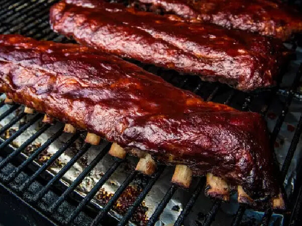 Two racks of ribs coated in barbecue sauce cooking on a grill with visible grill grates and a smoky background.