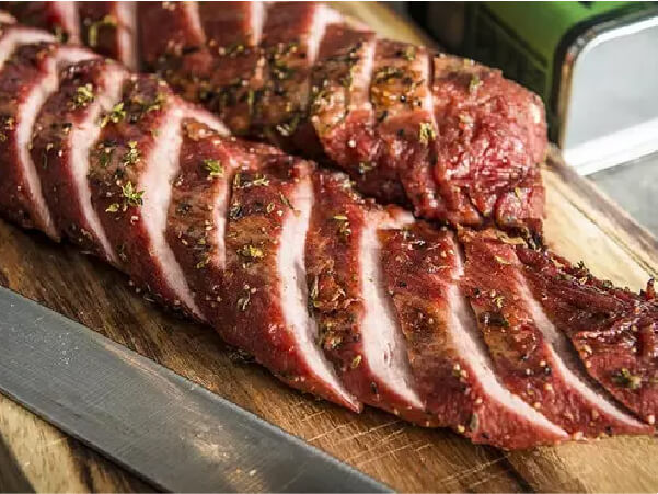 Sliced, cooked pork tenderloin with seasoning on a wooden cutting board next to a knife and a green container in the background.