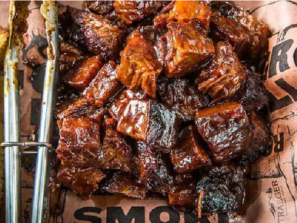 Close-up of bite-sized smoked beef chunks with a glossy caramelized exterior resting on branded butcher paper next to metal tongs.