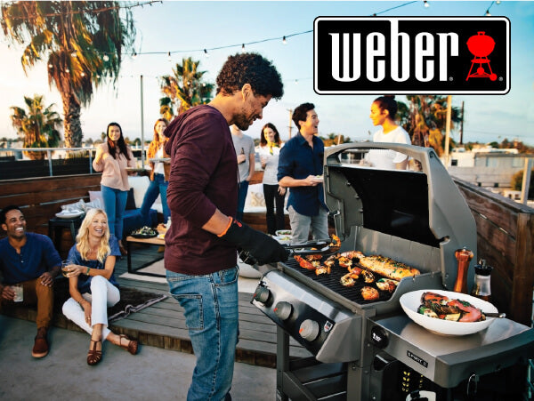 Man cooking various grilled foods on a gas barbecue grill outdoors while friends gather and chat on a rooftop patio at sunset.